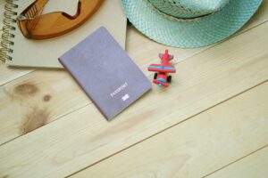 Flat lay of travel essentials including passport, notebook, toy airplane, sunglasses, and straw hat.